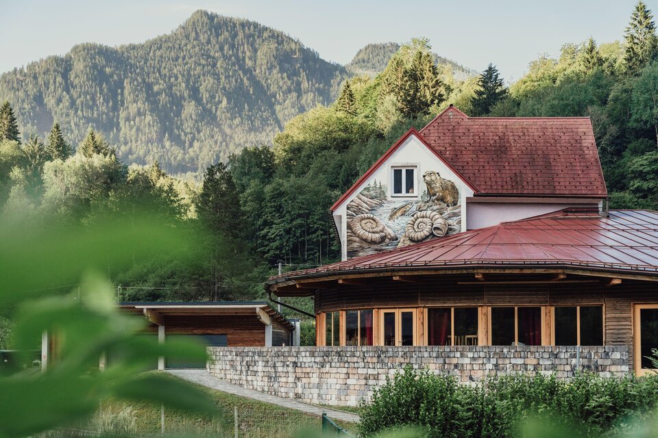 Ein charmantes Gebäude mit traditioneller Architektur liegt in einer malerischen Berglandschaft. Grüne Bäume und sanfte Hügel umgeben die Szene.