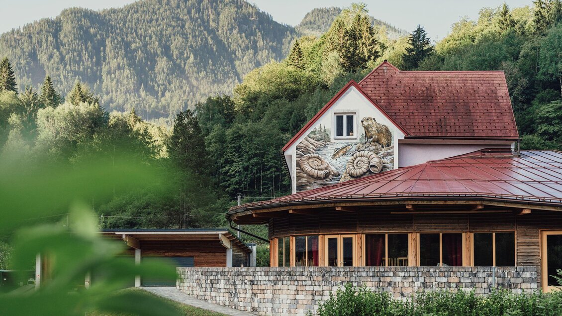 Ein charmantes Gebäude mit traditioneller Architektur liegt in einer malerischen Berglandschaft. Grüne Bäume und sanfte Hügel umgeben die Szene.