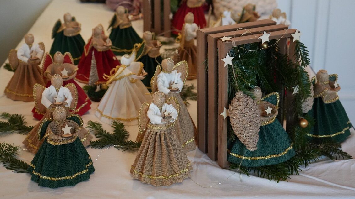 Eine Sammlung von handgemachten Engeln in festlichen Kleidern aus Stoff. Die Engel stehen auf einem Tisch und sind von grünen Zweigen und Weihnachtsdekoration umgeben. | © Peter Haslebner