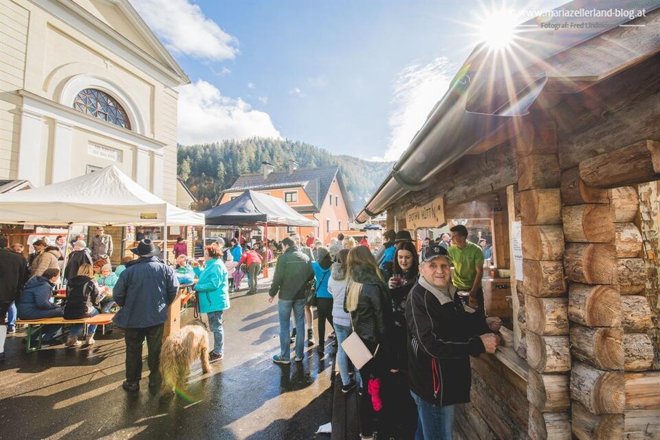 Ein belebter Markt mit vielen Menschen und Ständen. Die Sonne scheint über einer malerischen Landschaft. | © www.mariazell.blog