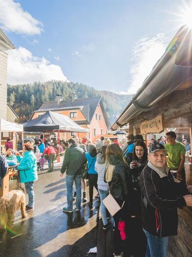 Ein belebter Markt mit vielen Menschen und Ständen. Die Sonne scheint über einer malerischen Landschaft. | © www.mariazell.blog