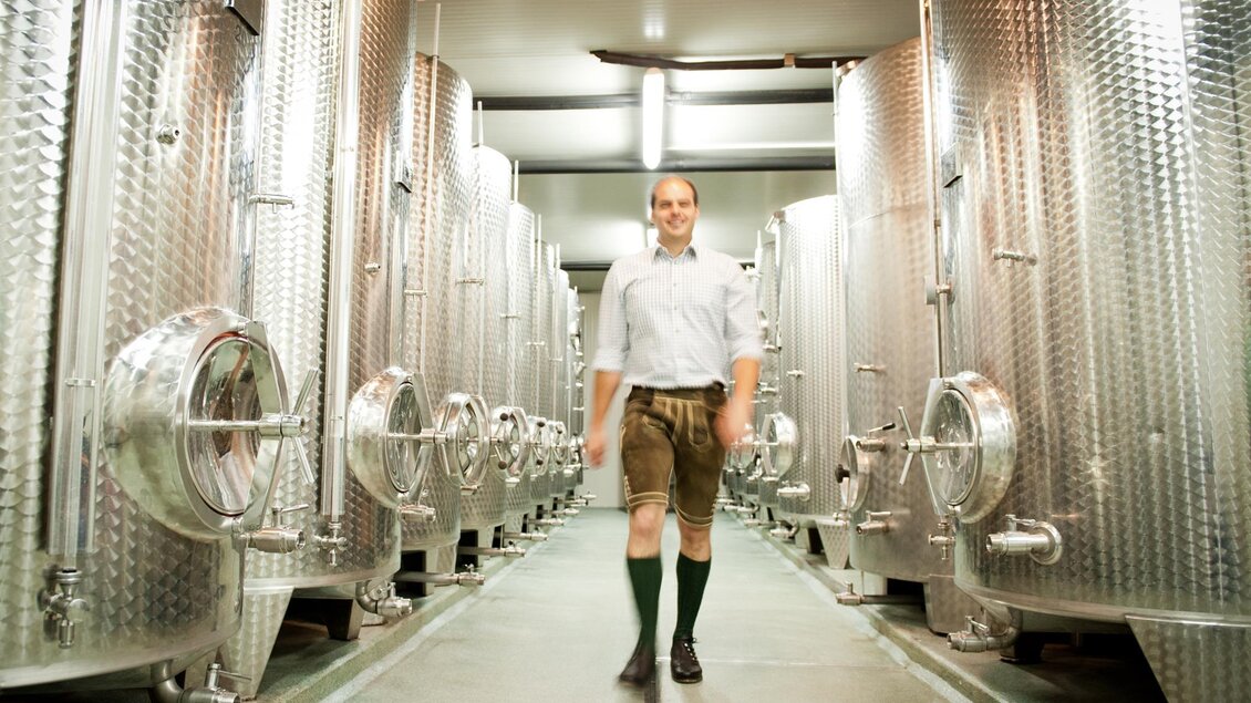 Ein Mann in traditioneller Tracht steht in einem Weinkeller mit großen Edelstahlbehältern. Der Raum wirkt modern und gut beleuchtet. | © Buschenschank Tinnauer