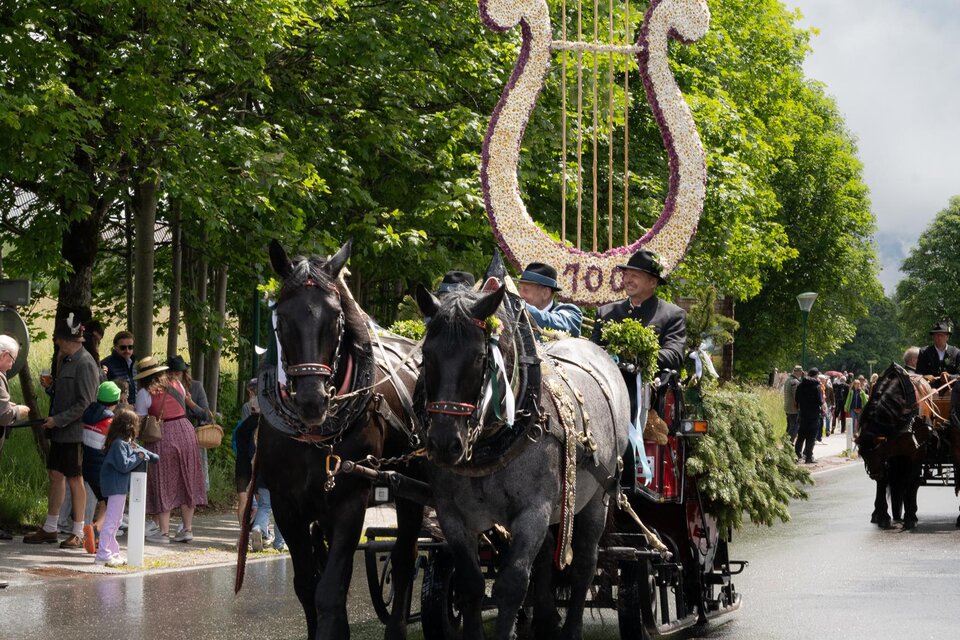 Eine Pferdekutsche mit einem dekorativen, blühenden Symbol in Form einer Harfe fährt eine Straße entlang. Umgeben von grünen Bäumen stehen Zuschauer am Straßenrand und beobachten die Parade.
