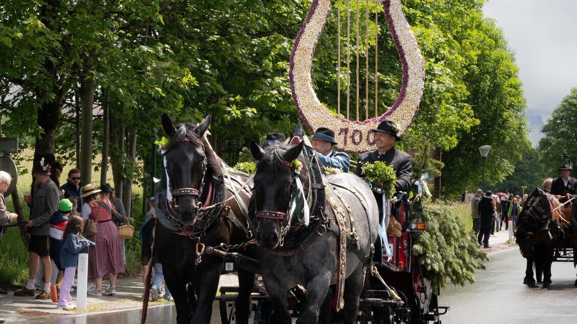 Eine Pferdekutsche mit einem dekorativen, blühenden Symbol in Form einer Harfe fährt eine Straße entlang. Umgeben von grünen Bäumen stehen Zuschauer am Straßenrand und beobachten die Parade.