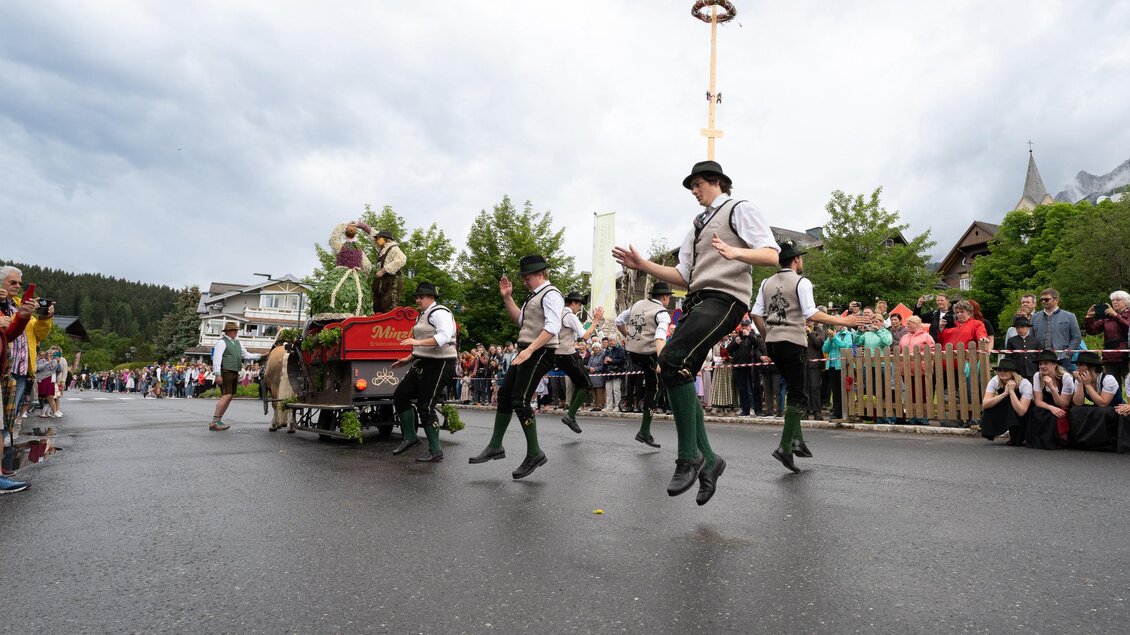 Eine traditionelle Festveranstaltung mit tanzenden Personen in Trachten. Im Hintergrund sind Zuschauer und eine festliche Atmosphäre sichtbar.