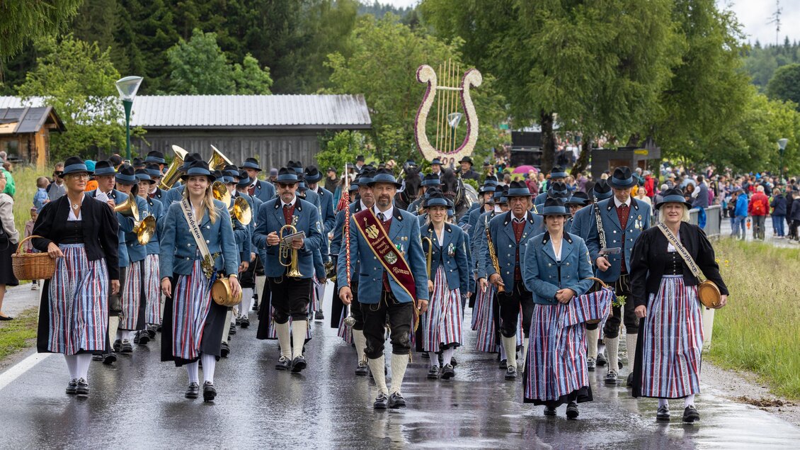 Eine Marching Band in traditionellen Trachten zieht durch eine Straße. Im Hintergrund sind Zuschauer und eine grüne Landschaft zu sehen.
