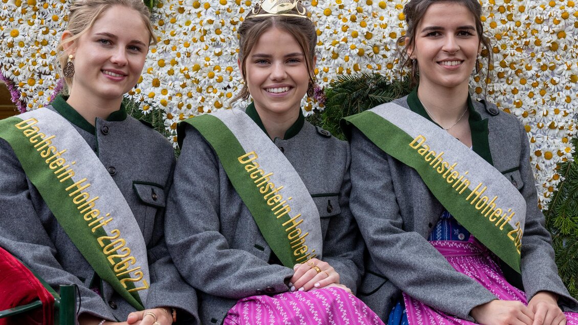 Drei Frauen in traditionellen Trachten sitzen lächelnd auf einem Wagen, umgeben von einer blühenden Dekoration. Sie tragen Gürtel mit Aufschriften und haben Kränze auf den Köpfen.