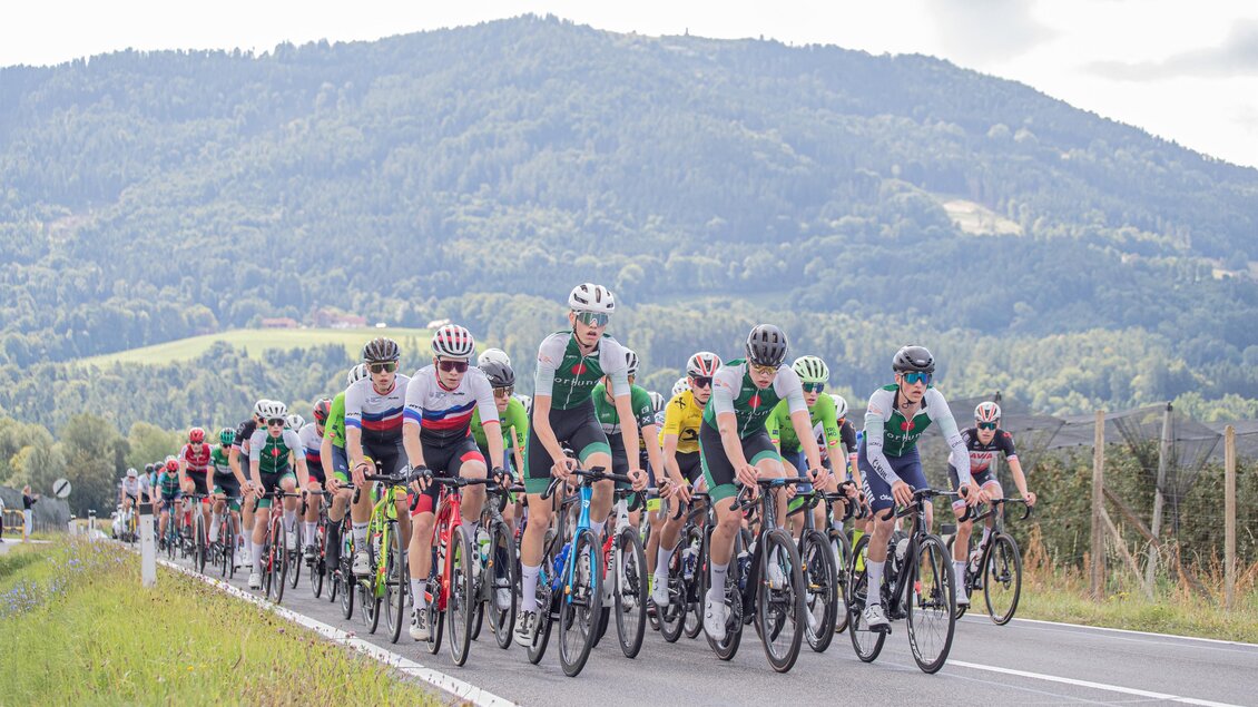Eine Gruppe von Radfahrern fährt auf einer Landstraße. Im Hintergrund sind grüne Berge und ein bewölkter Himmel zu sehen. | © Oststeiermark Tourismnus
