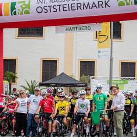 Eine Gruppe von Radfahrern steht unter einem Banner, das für die INT. Radjugendtour Oststeiermark wirbt. Im Hintergrund sind einige Zuschauer und Veranstalter zu sehen. | © Cycling Austria