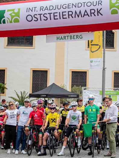 Eine Gruppe von Radfahrern steht unter einem Banner, das für die INT. Radjugendtour Oststeiermark wirbt. Im Hintergrund sind einige Zuschauer und Veranstalter zu sehen. | © Cycling Austria