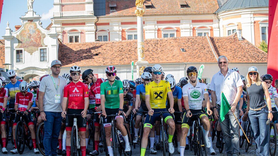 Eine Gruppe von Radfahrern steht an der Startlinie eines Rennens in Pollau. Im Hintergrund sind historische Gebäude und eine festliche Atmosphäre zu sehen. | © Peter Maurer