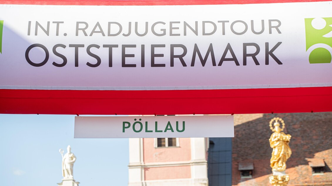 Ein Radrennen startet unter einem bunten Bogen in der Stadt Pollau. Im Hintergrund sind historische Gebäude und eine klare blaue Himmel zu sehen. | © Tourismusverband Oststeiermark