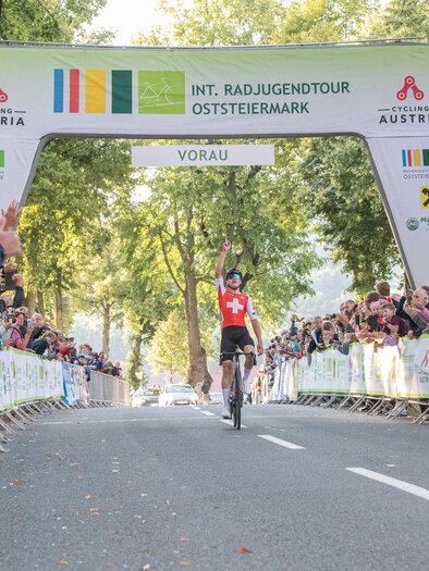 Ein Radsportler fährt unter einem Zielbogen hindurch, während die Menge jubelt. Es ist eine Veranstaltung der Int. Radsporttour in der Oststeiermark. | © Cycling Austria_Peter Maurer