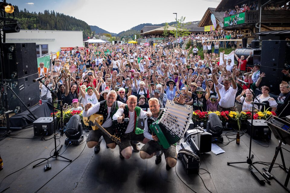 Eine lebhafte Veranstaltung mit einer großen Menschenmenge, die fröhlich feiert. Im Vordergrund stehen drei Musiker, die mit ihren Instrumenten posieren. | © Edlseer Alm BePo