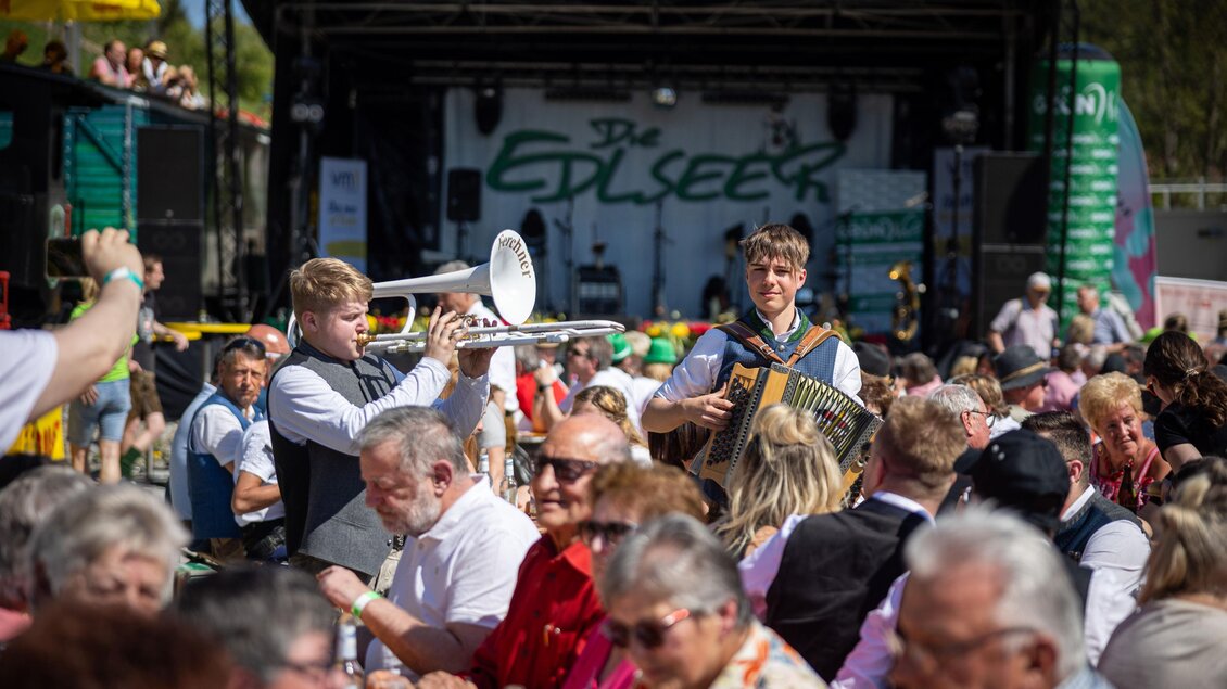 Ein lebhaftes Festival mit einer Band auf der Bühne. Eine Gruppe von Menschen sitzt an Tischen und genießt die Musik und das Ambiente. | © Edlseer Alm BePo