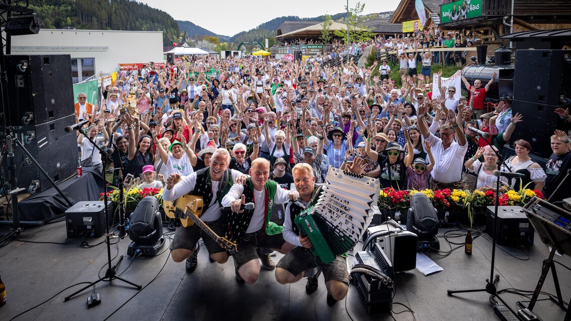 Eine lebhafte Veranstaltung mit einer großen Menschenmenge, die fröhlich feiert. Im Vordergrund stehen drei Musiker, die mit ihren Instrumenten posieren. | © Edlseer Alm BePo