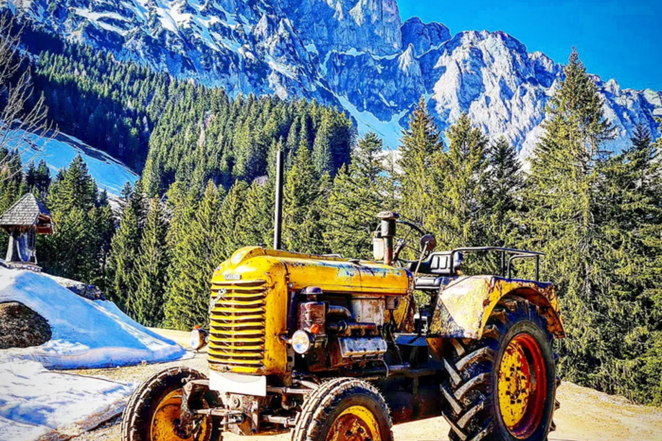 Ein gelber Traktor steht auf einem Weg in den Bergen. Im Hintergrund sind schneebedeckte Gipfel und grüne Wälder zu sehen. | © Brandmüller Lorenz