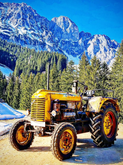 Ein gelber Traktor steht auf einem Weg in den Bergen. Im Hintergrund sind schneebedeckte Gipfel und grüne Wälder zu sehen. | © Brandmüller Lorenz