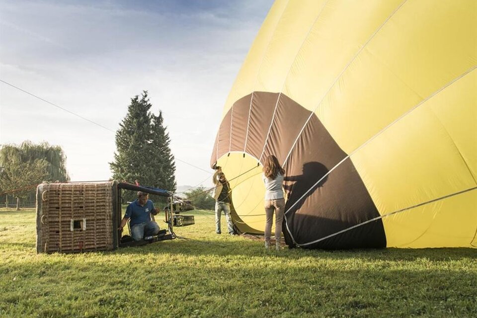 Ein Heißluftballon wird aufgebaut, während die Crew zusammenarbeitet. Die Szene findet auf einem grünen Feld unter blauem Himmel statt. | © Marion Luttenberger