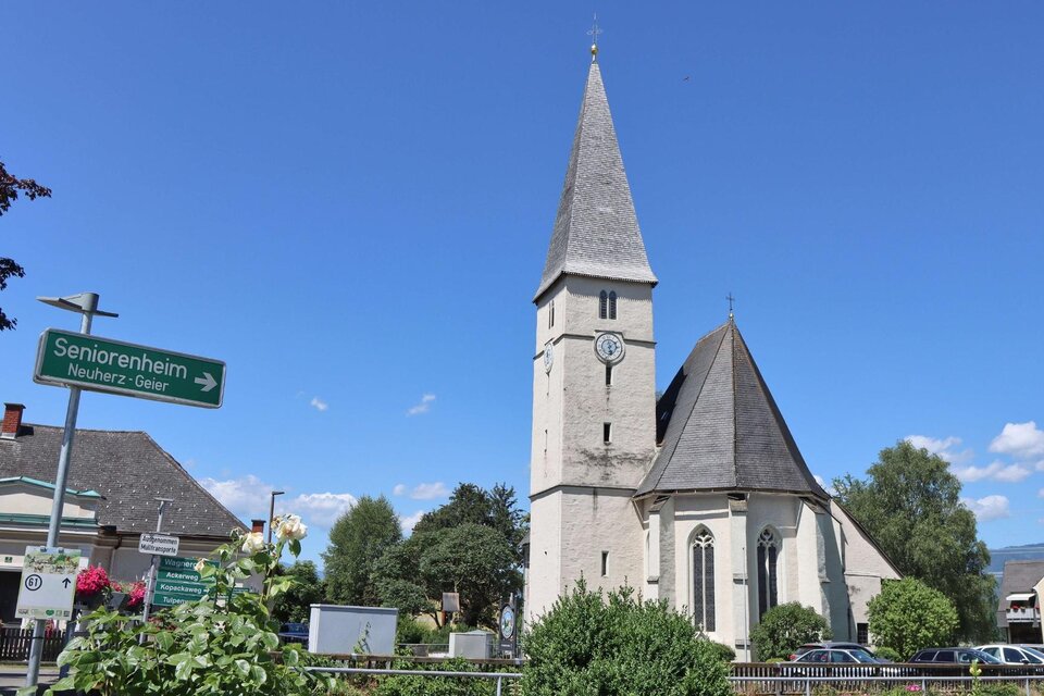 Eine schöne Kirche mit einem hohen Turm steht in einem kleinen Dorf. Im Vordergrund sind grüne Sträucher und ein Wegweiser zu sehen. | © Anita Fössl