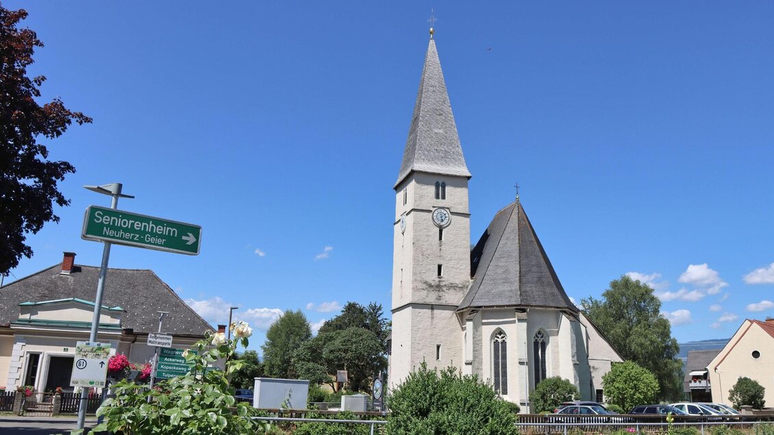 Eine schöne Kirche mit einem hohen Turm steht in einem kleinen Dorf. Im Vordergrund sind grüne Sträucher und ein Wegweiser zu sehen. | © Anita Fössl