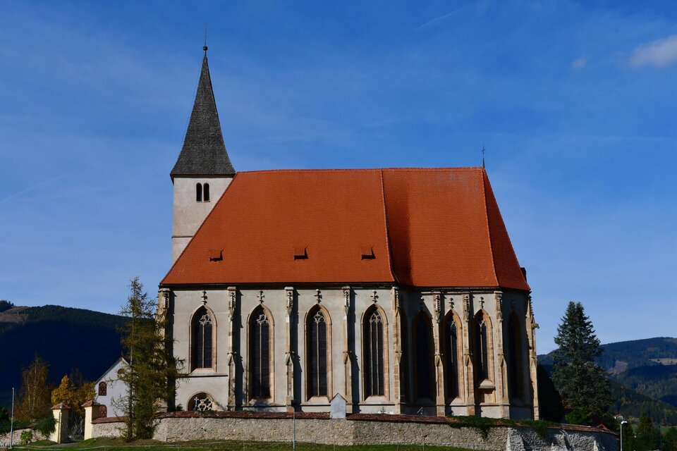 Eine hübsche Kirche mit einem roten Dach und hohen Fenstern. Im Hintergrund sind sanfte Hügel und ein klarer Himmel zu sehen. | © Josef Reibenbacher