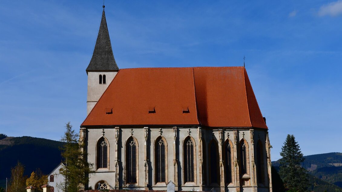 Eine hübsche Kirche mit einem roten Dach und hohen Fenstern. Im Hintergrund sind sanfte Hügel und ein klarer Himmel zu sehen. | © Josef Reibenbacher
