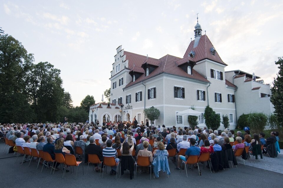 Eine große Menschenmenge sitzt vor einem Schloss. Im Hintergrund ist die Architektur des Gebäudes und der Abendhimmel zu sehen. | © Lore Schrettner
