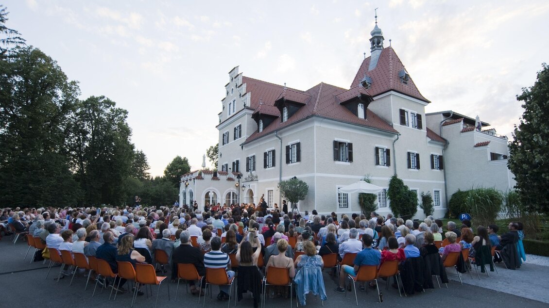 Eine große Menschenmenge sitzt vor einem Schloss. Im Hintergrund ist die Architektur des Gebäudes und der Abendhimmel zu sehen. | © Lore Schrettner