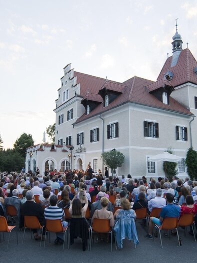 Eine große Menschenmenge sitzt vor einem Schloss. Im Hintergrund ist die Architektur des Gebäudes und der Abendhimmel zu sehen. | © Lore Schrettner