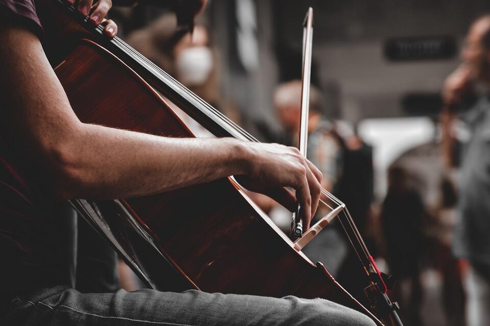 Eine Hand spielt auf einem Cello in einer belebten Umgebung. Im Hintergrund sind unscharfe Menschen zu sehen. | © pixabay.com