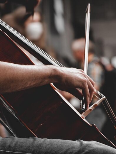Eine Hand spielt auf einem Cello in einer belebten Umgebung. Im Hintergrund sind unscharfe Menschen zu sehen. | © pixabay.com