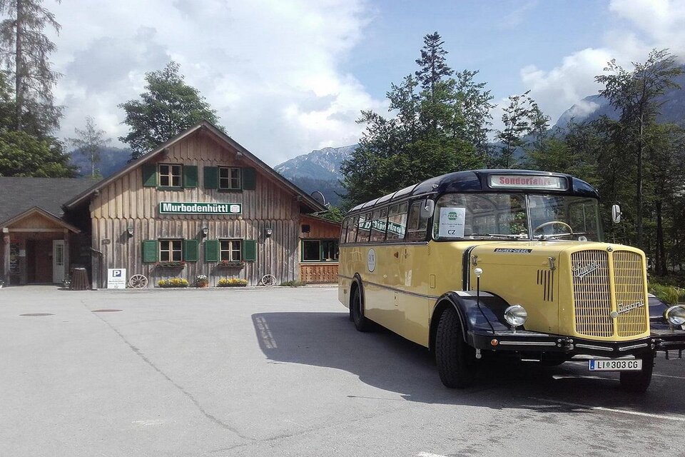 Ein gelber Oldtimer-Bus steht vor einem traditionellen Holzhaus in einer malerischen Landschaft. Im Hintergrund sind Berge und Bäume sichtbar. | © Peter Prommer