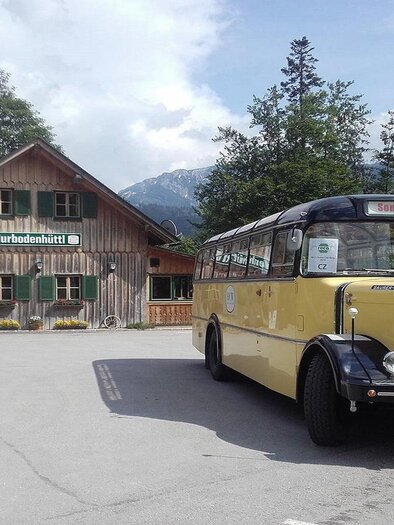 A yellow vintage bus is standing in front of a traditional wooden house in a picturesque landscape. In the background, mountains and trees are visible. | © Peter Prommer