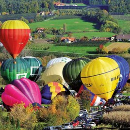 Bunte Heißluftballons schweben über einer malerischen Landschaft mit sanften Hügeln und vielen Bäumen. Darunter parken Autos, während die Wiesen in herbstlichen Farben leuchten. | © Zugschwert