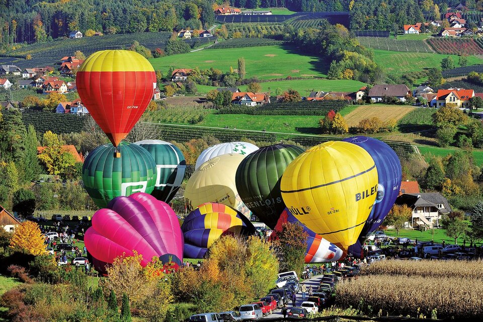 Bunte Heißluftballons schweben über einer malerischen Landschaft mit sanften Hügeln und vielen Bäumen. Darunter parken Autos, während die Wiesen in herbstlichen Farben leuchten. | © Zugschwert