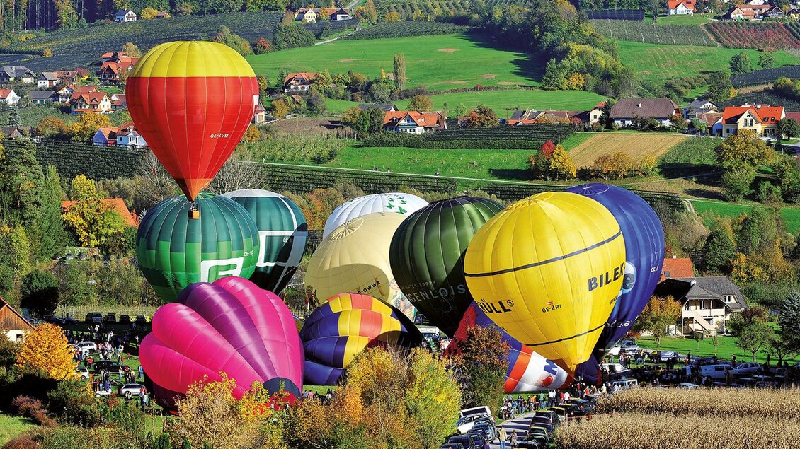 Bunte Heißluftballons schweben über einer malerischen Landschaft mit sanften Hügeln und vielen Bäumen. Darunter parken Autos, während die Wiesen in herbstlichen Farben leuchten. | © Zugschwert