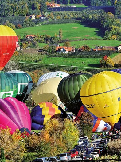 Bunte Heißluftballons schweben über einer malerischen Landschaft mit sanften Hügeln und vielen Bäumen. Darunter parken Autos, während die Wiesen in herbstlichen Farben leuchten. | © Zugschwert