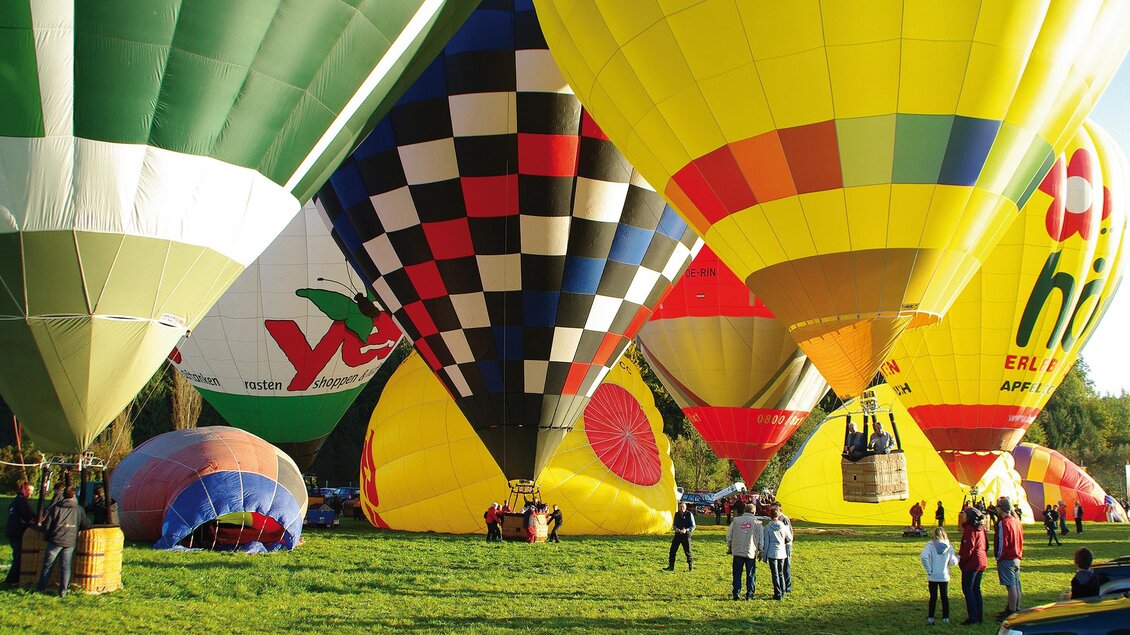Bunte Heißluftballons stehen auf einer grünen Wiese. Menschen beobachten die Vorbereitungen zum Start. | © Haring