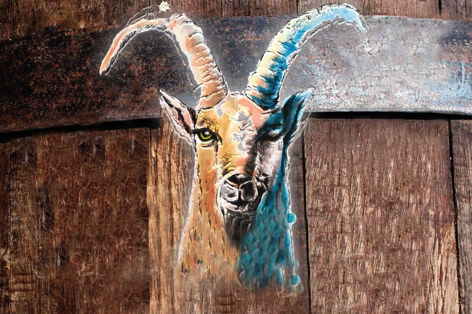 Ein kunstvolles Bild eines Ziegenkopfes mit großen Hörnern, das auf einem holzernen Hintergrund gemalt ist. Die Farben sind lebhaft und betonen die Details des Gesichts.