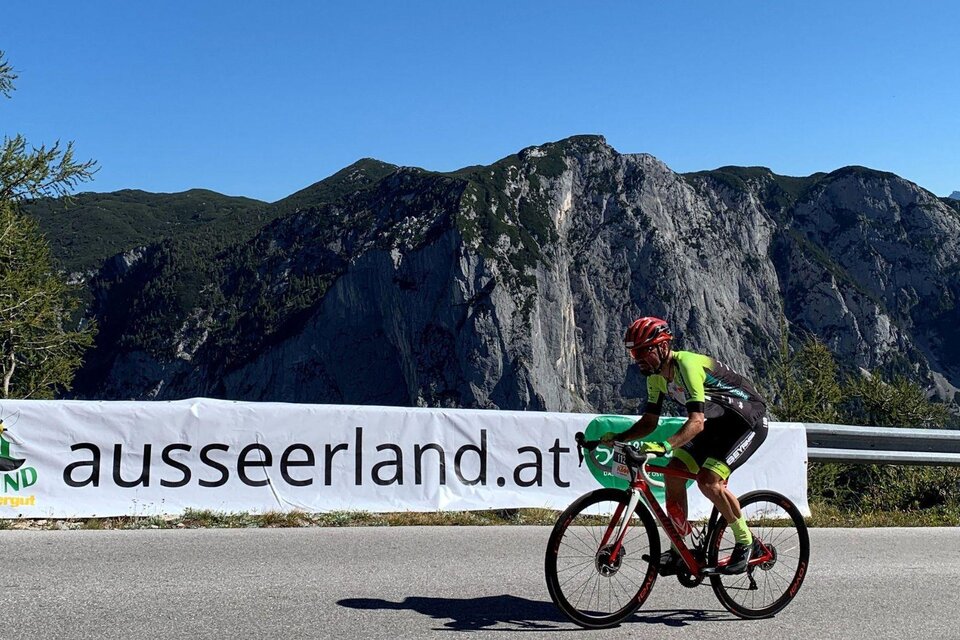 Ein Radfahrer fährt auf einer Straße mit bergiger Kulisse im Hintergrund. Im Vordergrund befindet sich ein Banner mit der Aufschrift "ausseerland.at". | © Lindpointner