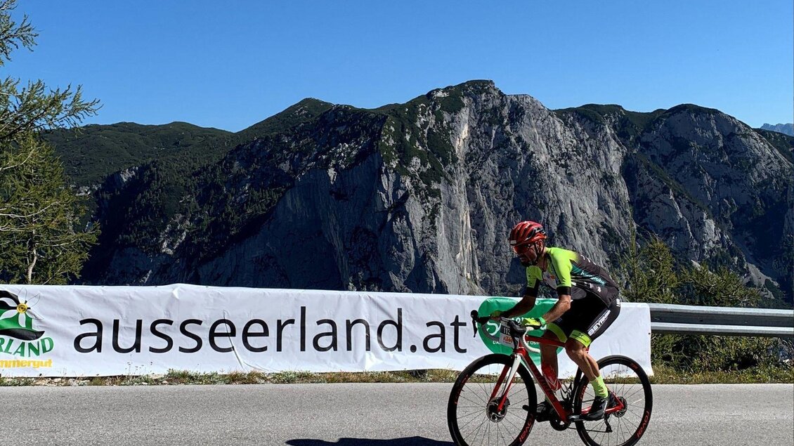 Ein Radfahrer fährt auf einer Straße mit bergiger Kulisse im Hintergrund. Im Vordergrund befindet sich ein Banner mit der Aufschrift "ausseerland.at". | © Lindpointner