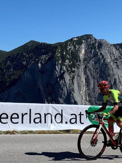 A cyclist rides on a road with a hilly backdrop in the background. In the foreground, there is a banner with the inscription "ausseerland.at". | © Lindpointner