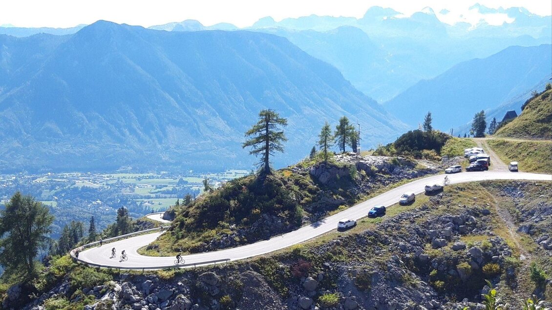 Eine kurvenreiche Straße in den Bergen mit Radfahrern und Autos. Im Hintergrund sind hohe Berge und eine grüne Landschaft zu sehen. | © Peter Perstl