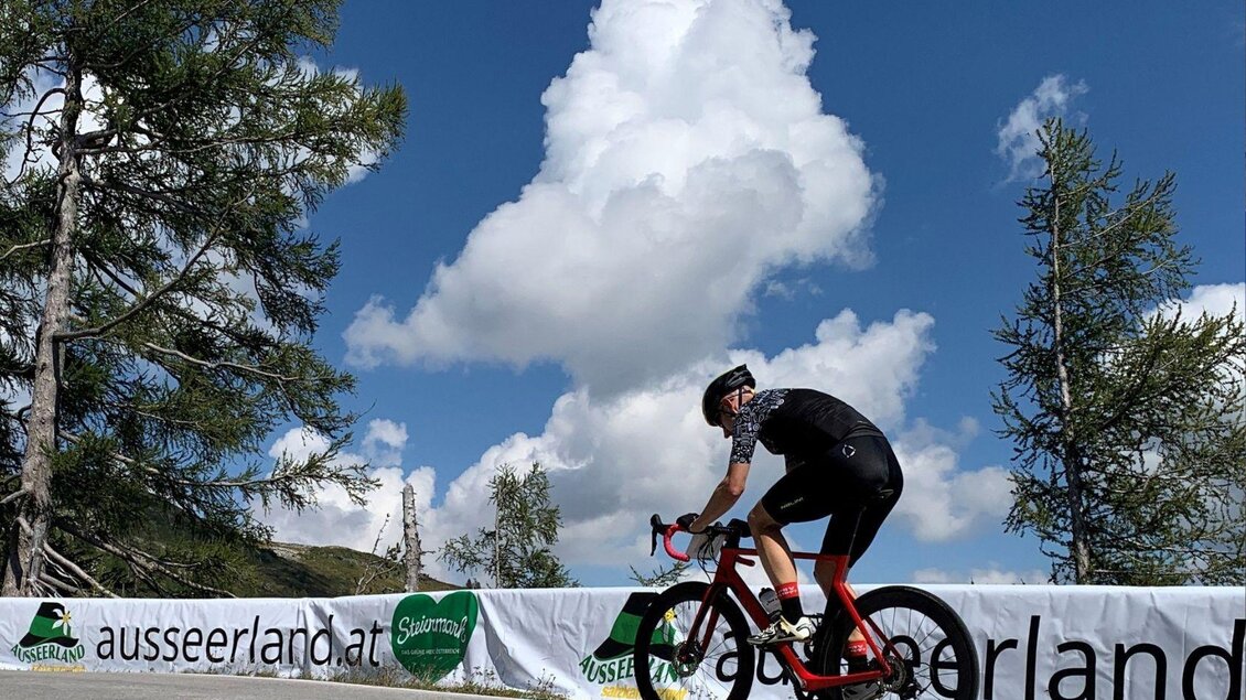Ein Radfahrer fährt entlang einer Straße unter einem klaren Himmel mit großen Wolken. Im Hintergrund sind Bäume und eine Werbebande zu sehen. | © Lindpointner