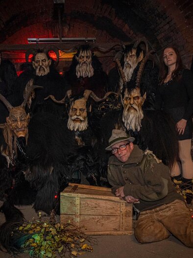 Eine Gruppe von Personen in einer dunklen Umgebung, gekleidet in schaurige Kostüme mit Tiermasken und schwarzen Flügeln. Im Vordergrund sitzt eine Person auf dem Boden neben einer Holztruhe. | © Vordernberger Hunt's Teufel