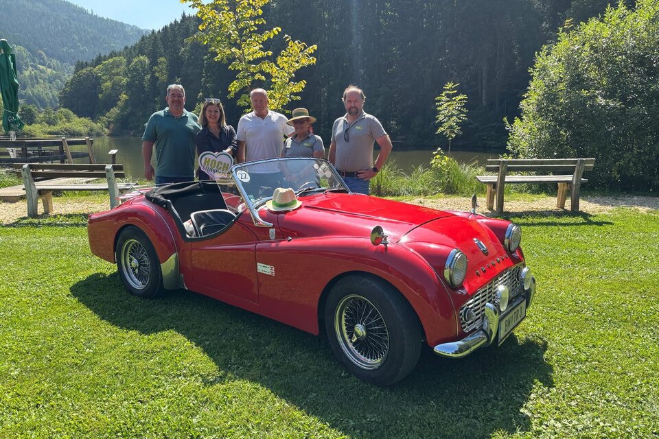 Eine Gruppe von fünf Personen steht neben einem roten Sportwagen auf einer grünen Wiese. Im Hintergrund sind Bäume und ein ruhiger See zu sehen.