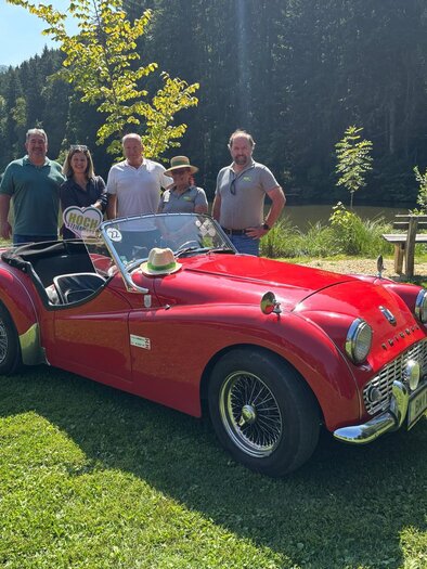 A group of five people stands next to a red sports car on a green meadow. In the background, there are trees and a calm lake.