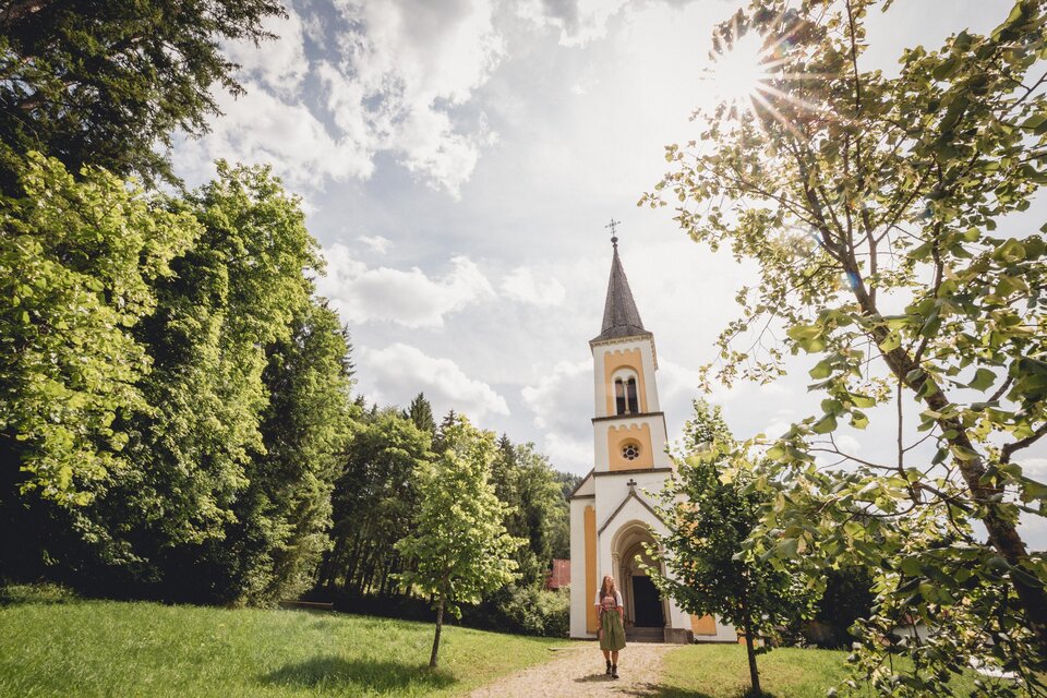 Eine malerische Kirche steht unter einem klaren Himmel mit Sonne. Der Weg zur Kirche ist von grünen Bäumen gesäumt. | © TV Hochsteiermark, nicoleseiser.at