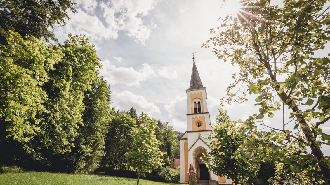 Eine malerische Kirche steht unter einem klaren Himmel mit Sonne. Der Weg zur Kirche ist von grünen Bäumen gesäumt. | © TV Hochsteiermark, nicoleseiser.at